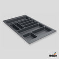 A dark gray plastic under-sink organizer tray with multiple compartments, shown at an angle on a light gray background with the Hettich brand logo in the bottom right corner.